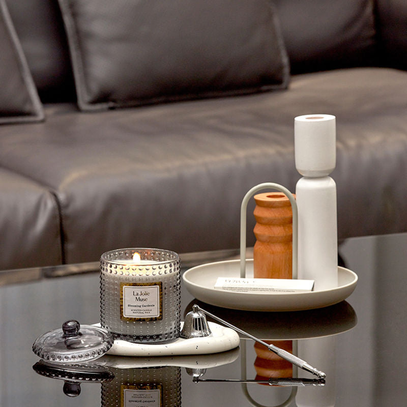 A burning candle and a candle snuffer are on a glass top table in front of a brown coach. 