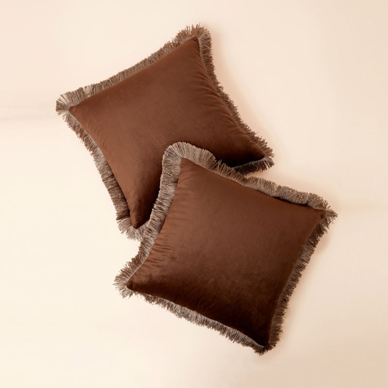 A full view of a set of two 18 * 18 inch bronze tassel fringed pillows in cover is displayed in a staggered position.