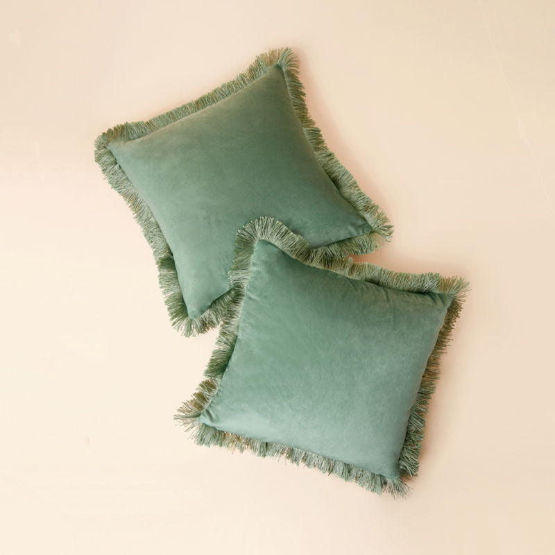 A full view of a set of two 18 * 18 inch green tassel fringed pillows in cover is displayed in a staggered position.