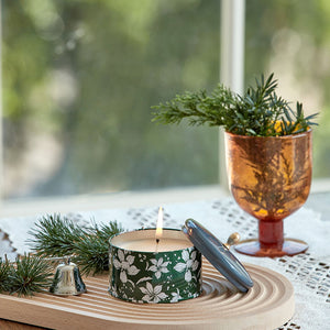 A jar of burning candle is placed on a wooden tray, surrounded by pine needles and a glass of pine branches.
