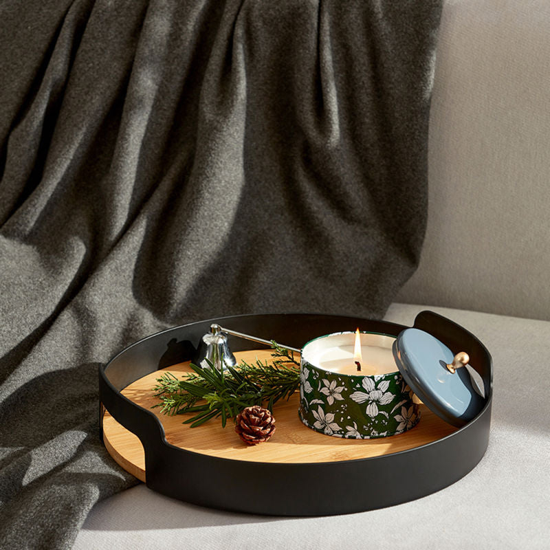 A burning candle is placed in a black woody tray with pine branches and a pine cone.