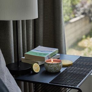 A burning candle is placed on a bedside table next to a window, with two books and a cut green lime beside.