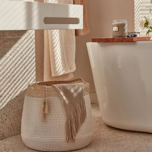 The cotton and corn skin hamper is placed next to a bathtub, the shadow of shutters on the marble wall in the background.