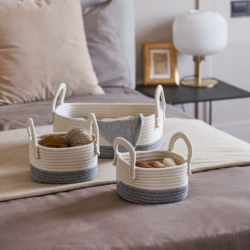 Three cotton rope baskets storing balls of yarn are displayed on a bed. A bedside table with a lamp is in the back.