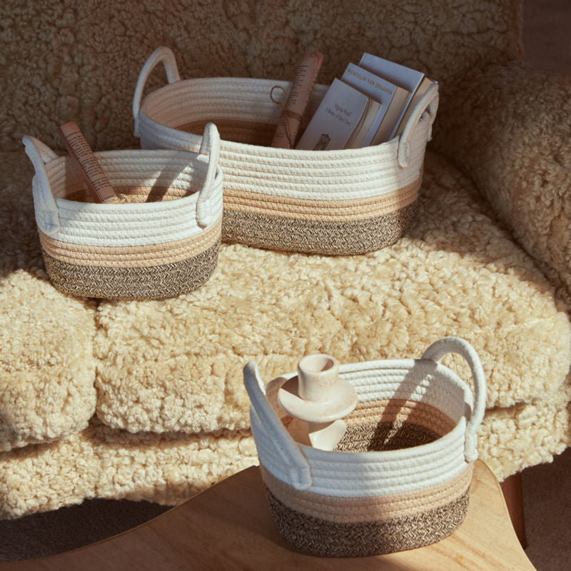 Three cotton rope baskets filled up with books are placed o a fluffy sofa, under the sunshine.