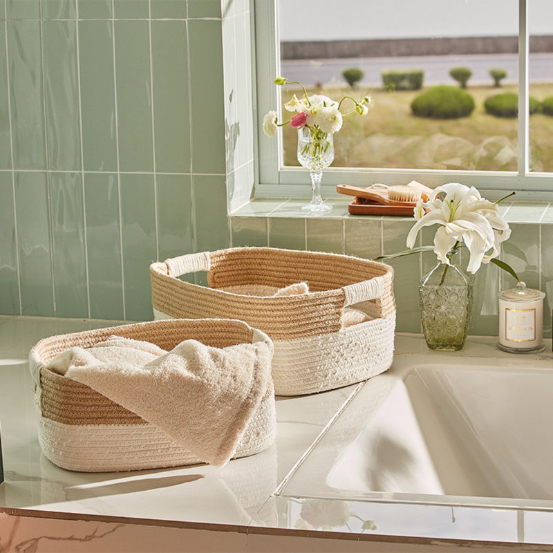 Two cotton rope and jute baskets are placed next to a bathtub, storing towels inside. Surrounded by a candle and vases of lilies.