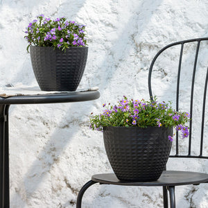 Two black planters with cube pattern are displayed in front of a granite textured wall, both potted with purple flowers.