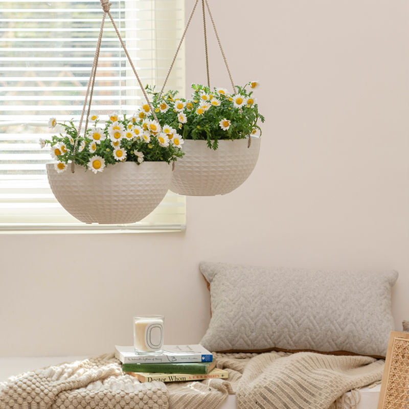 A pair of matte white planters are hung in front of white blinds, each potted with white flowers.