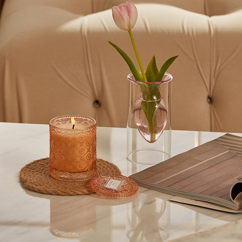 A burning candle is placed on a white marble table with a rattan cup pad underneath, accompanied by a vase of tulip.