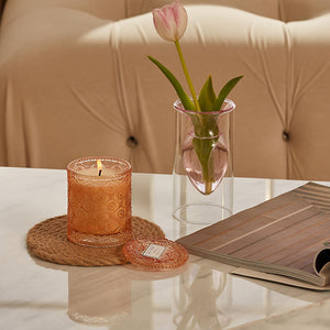 A burning candle is placed on a white marble table with a rattan cup pad underneath, accompanied by a vase of tulip.