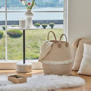 A beige laundry basket is surrounded by cushions and carpet, in front of a garden view outside the window.