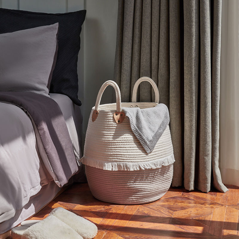 The large bulging laundry basket is displayed next to a bed, standing under the shadow of sunshine in the corner.
