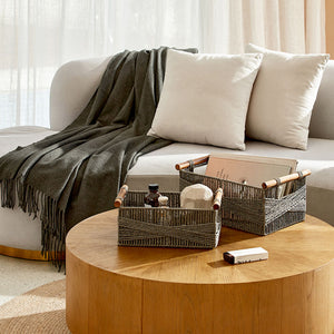 A set of two gray baskets are placed on a round wooden table, in front of a white sofa.