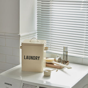 The beige laundry tin is displayed on a washing machine, surrounded by brushes and cleaning supplies.