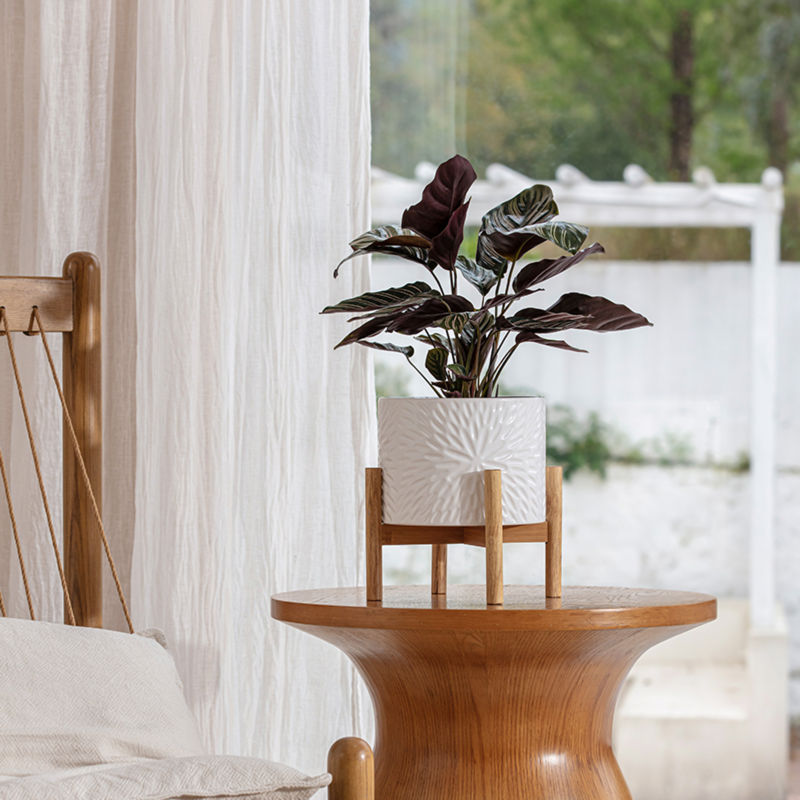 The white planter with its stand holds plants, is placed on a modern-designed wooden side table, next to a bed.