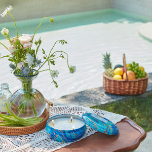A burning candle is placed on a small table by the swimming pool, with citronella and fruits as background.