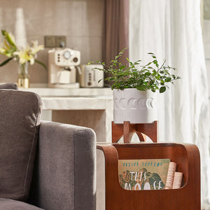 A white ceramic planter with a stand is placed on a small bookshelf, next to a gray sofa.