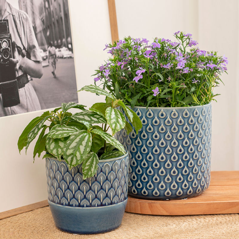 A set of two blue pots with flowers in them are placed on the ground, in front of a picture frame.