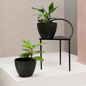 A set of two black outdoor planters are displayed in a staggered way, one on a black chair while the other one on the floor.