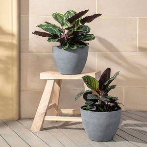 A set of two gray pots with plants in them are placed in front of the tile wall, one is on the chair and one is on the ground.