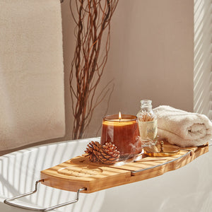 An oval jar of a burning candle is placed on a wooden board set across a white bathtub, accompanied by bath supplies.