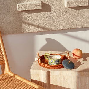 A jar of burning candle is displayed with fresh peach and kiwi fruit, next to a rattan chair.