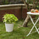 The matte white planter with plants in it is displayed on the outside ground, next to a white table.