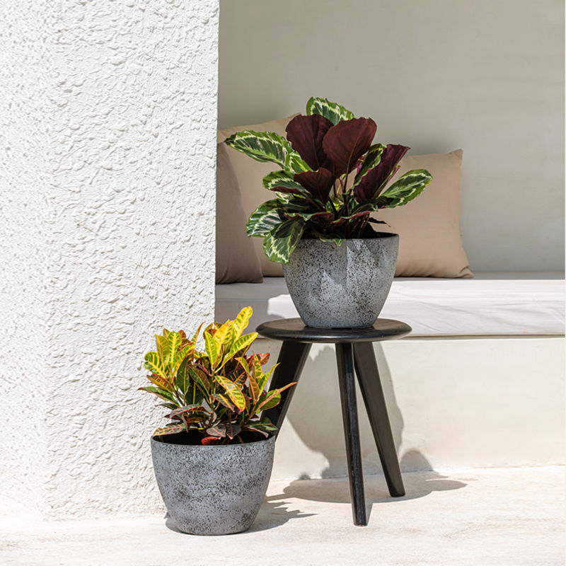 Two rock gray planters are displayed on a sunny patio, one on a small black chair and the other one is on the ground. 