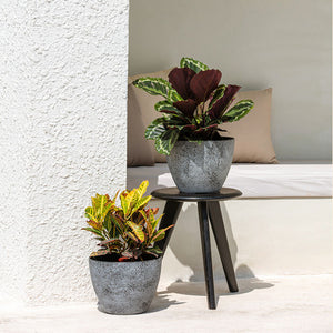 Two rock gray planters are displayed on a sunny patio, one on a small black chair and the other one is on the ground. 