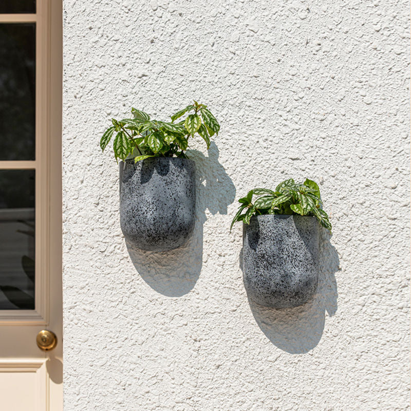 A pair of gray wall planters are hung outside, on a white wall.