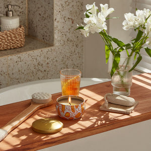 A burning candle is placed on a wooden board set across a bathtub, with flowers, bath supplies and a glass of juice beside.