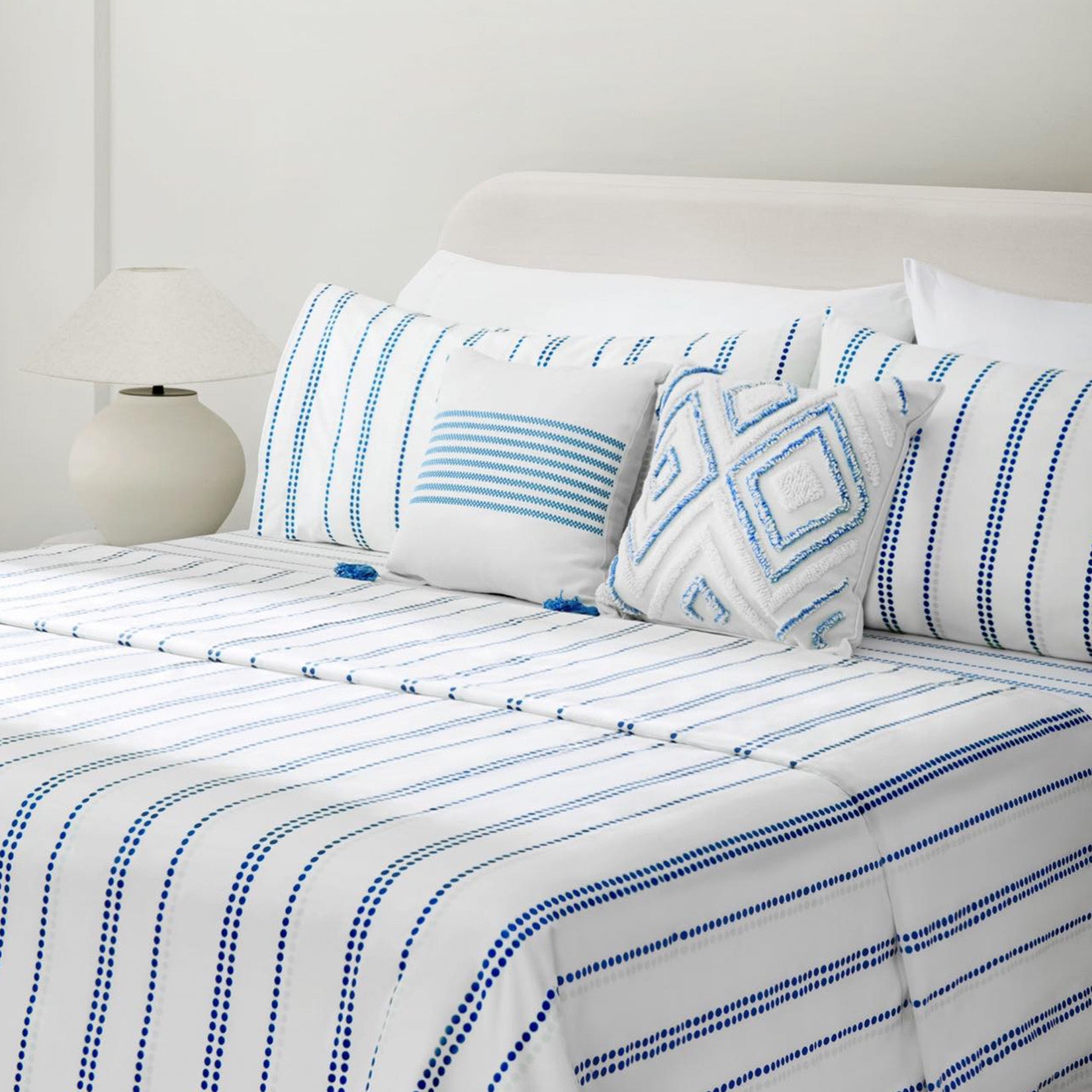 Camille Blue Stripe bed set. Blue and white dotted pillows and blue and white dotted sheets on bed with striped accent pillow and diamond tufted accent pillow with side table and lamp from side angle.