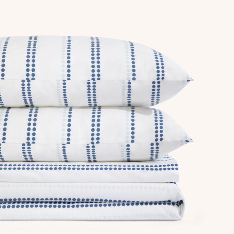 Camille Blue Stripe bed sheet set. Two blue and white dotted pillows stacked on folded blue and white dotted sheet set.