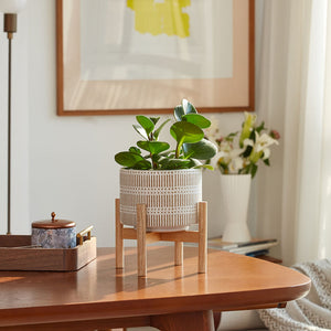 A sandy beige planter with base is placed on a wooden dining table, next to a wooden tray holding a candle jar. 