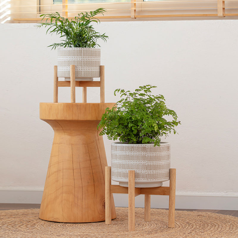 Two sandy beige ceramic planters are displayed in a staggered way, one on a small table and the other one on the floor. 