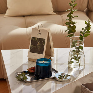 A burning candle is placed on a white marble table with a vase of eucalyptus branches and half a fig.