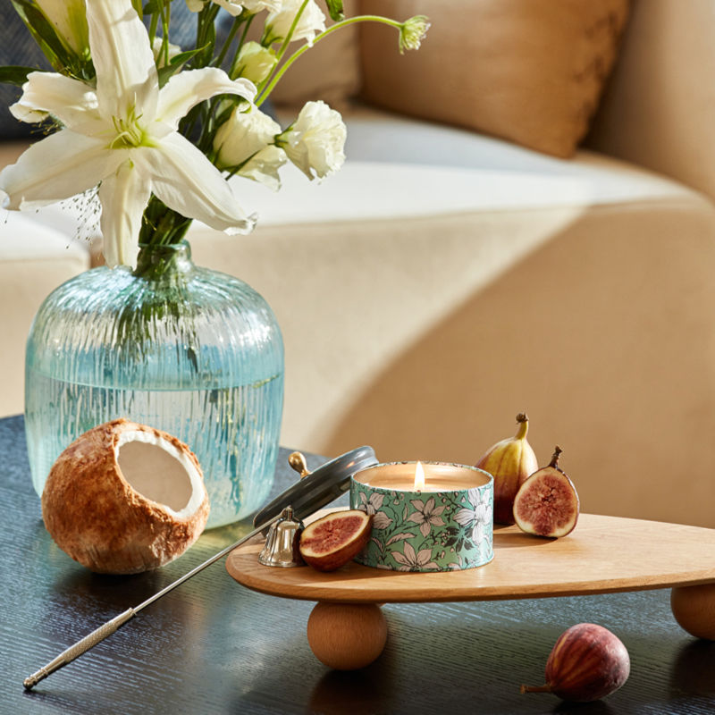 A burning candle is placed on a woody trolley tray with cut figs, in front of a opened coconut and a vase of white flowers.