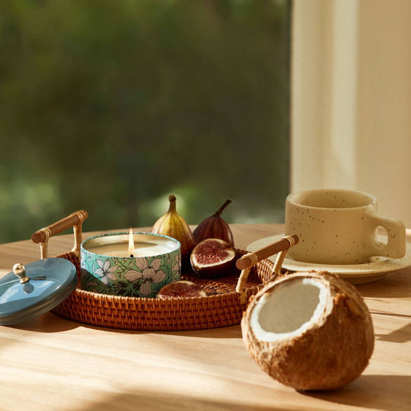 A burning candle is displayed in a rattan basket with some cut figs and a coconut.