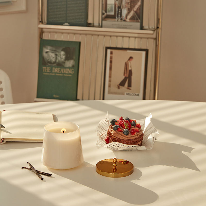 A white jar of burning candle is placed on a dining table with a delicious fruit cake.
