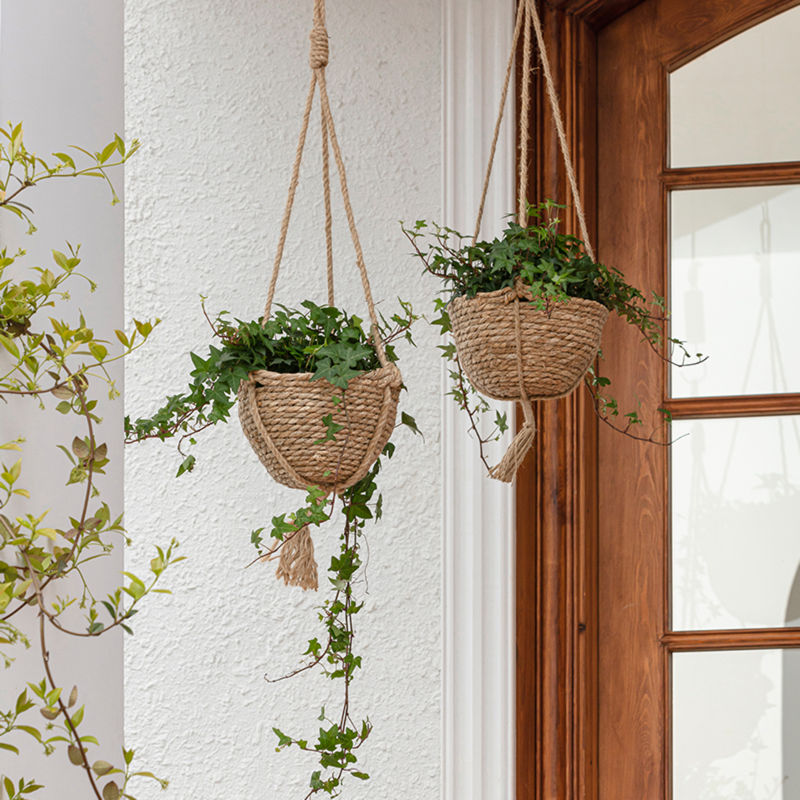 Two seagrass hanging planters are displayed in front of a wooden window, both potted with green plants.