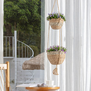 A set of two seagrass planters are hanging together in a space, in front of a window with opened curtain.