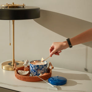 A hand is lighting a jar of candle on a lamp table next to a white wall.