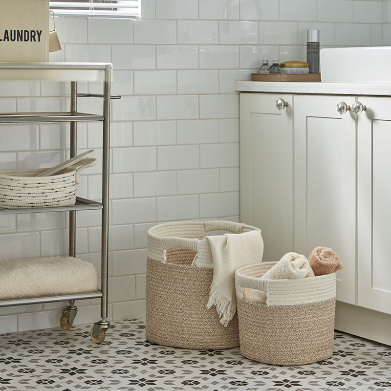 Two baskets in different sizes are placed on the tile floor, between a trolley and a white cabinet. 