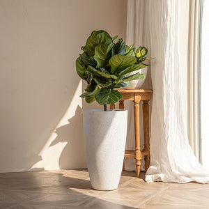 A speckled white tall planter is placed on the floor, in front of a white curtain.