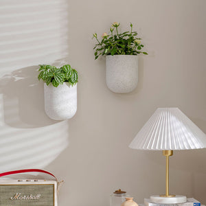 A pair of small planters are hung on a white wall, above a white table lamp.