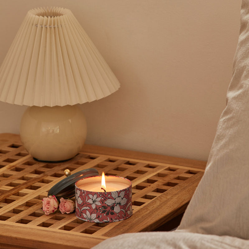A burning candle is placed on a wooden mesh bedside table, with a white table lamp behind.