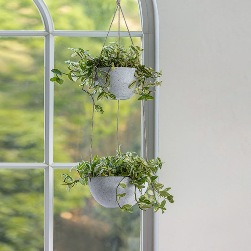 Two-tier white hanging planter is displayed in front of a window, potted with lush green plants.