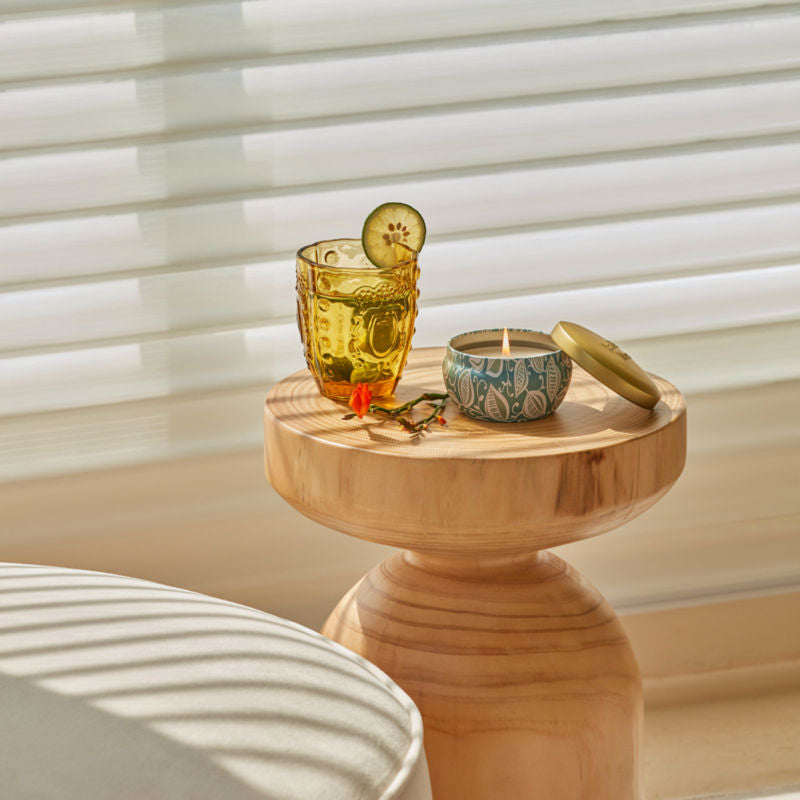 A burning candle is displayed on a small coffee table with a glass of orange flavored water, in front of white shutters.