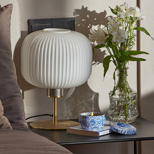 A burning candle is placed on a black bedside table with a white lamp and a vase of tulips.