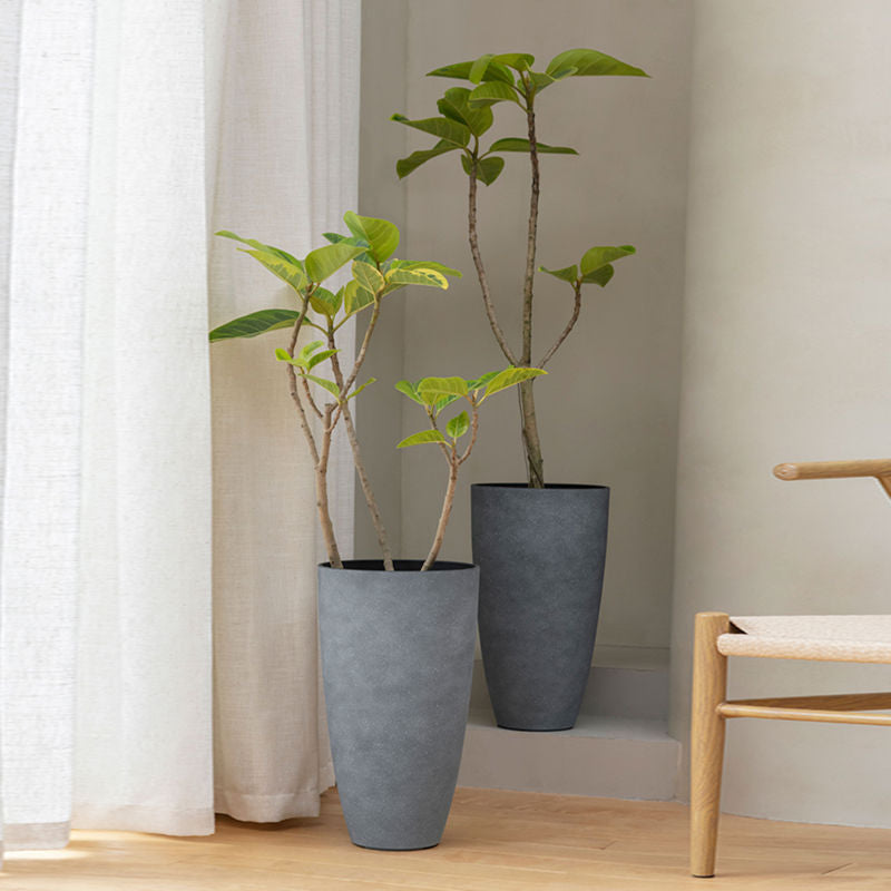 Two gray tall planters are displayed in a staggered way, between white curtains and a wooden chair.
